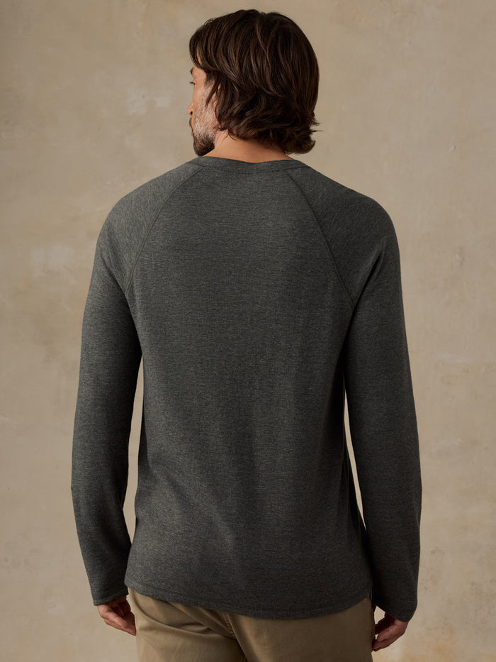 A man viewed from the back wearing a dark heather green long-sleeve raglan shirt, paired with tan pants, against a textured beige studio backdrop.
