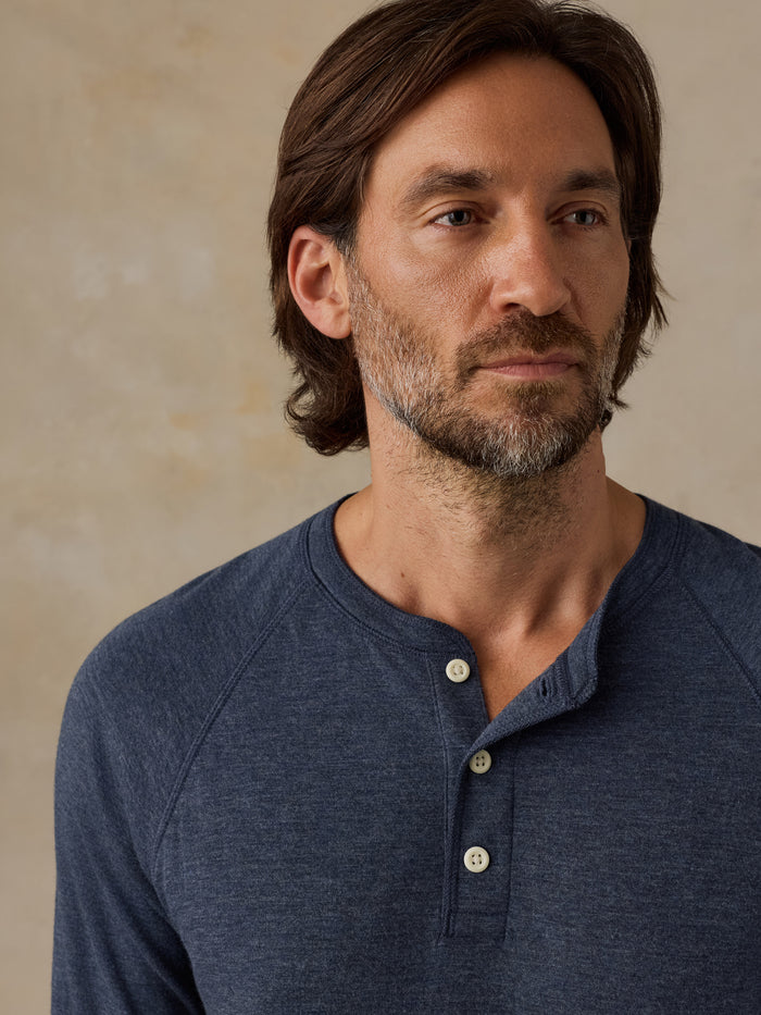 A man wearing a heathered navy blue henley shirt with three buttons, shown in a close-up portrait against a textured beige background.