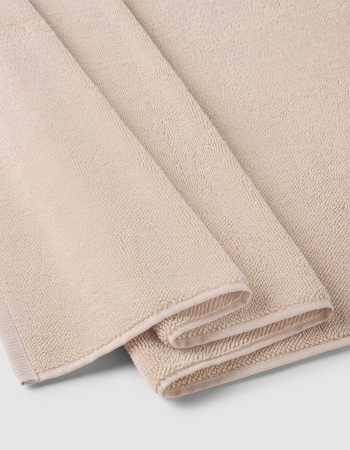 A Loop Terry Bath Mat by Cozy Earth, in light beige with a soft, textured surface, is neatly folded and stacked on a white background.|Color:Petal