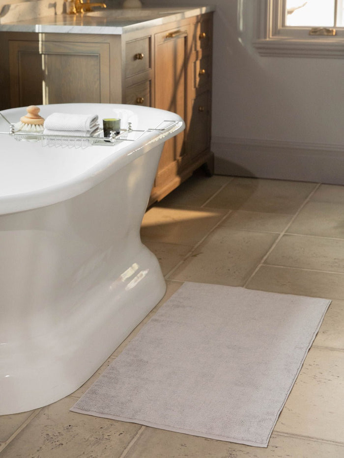 The Cozy Earth Loop Terry Bath Mat is shown on a tiled bathroom floor beside a freestanding tub, with folded towels and toiletries on a metal bath caddy. A wooden vanity with drawers appears in the background.|Color:Light Grey