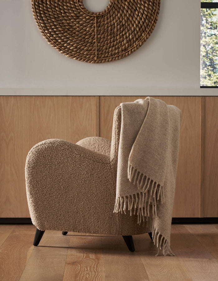A cozy beige textured armchair with a Cozy Earth Lucia Throw draped over the back sits on a wooden floor. Behind it are light wood cabinets and a round woven wall hanging, with sunlight streaming in through the window.|Color:Grain
