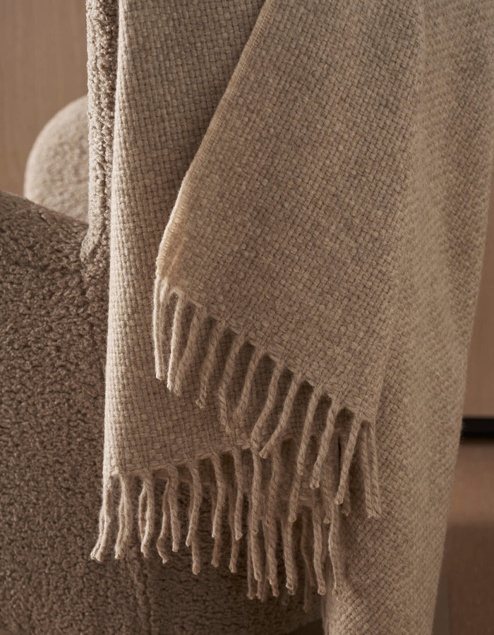 The Cozy Earth Lucia Throw, featuring a beige textured design with fringe, drapes over the arm of a soft upholstered chair in a neutral-toned room.|Color:Grain