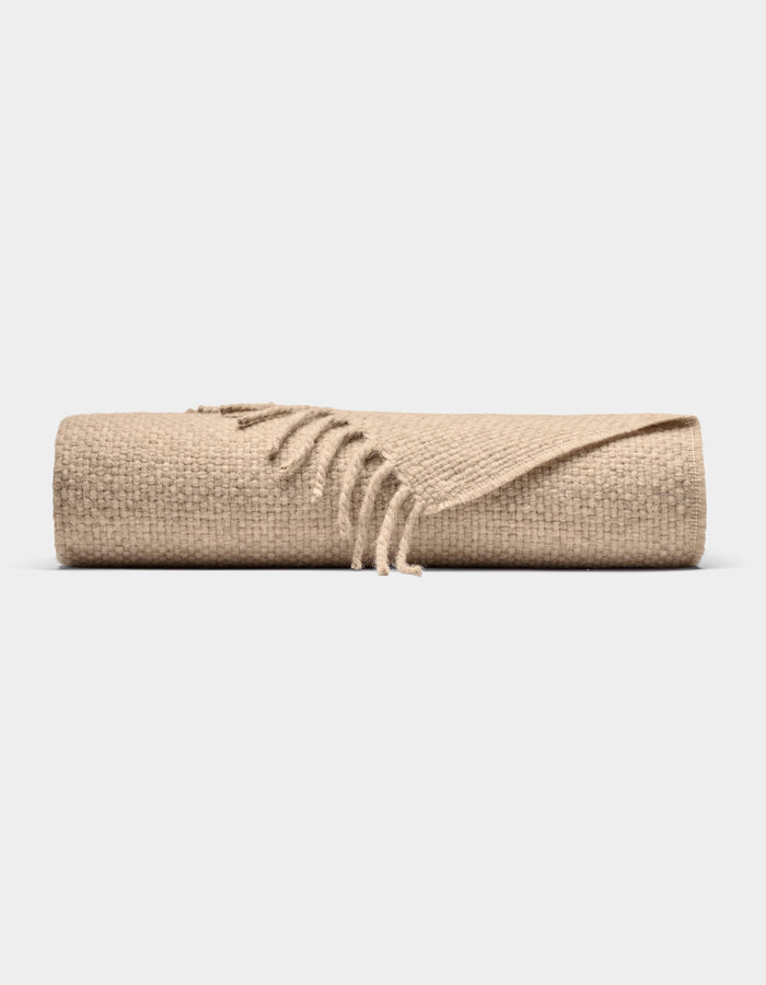 The Lucia Throw by Cozy Earth is a beige woven blanket with fringed edges, shown rolled up against a plain white background.|Color:Grain
