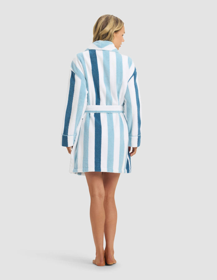 A woman with loose blonde hair stands barefoot, facing away, wearing the Cozy Earth Classic Stripe Resort Robe—knee-length with vertical blue and white stripes, a belted waist, against a plain light gray background.|Color:Lake Stripe