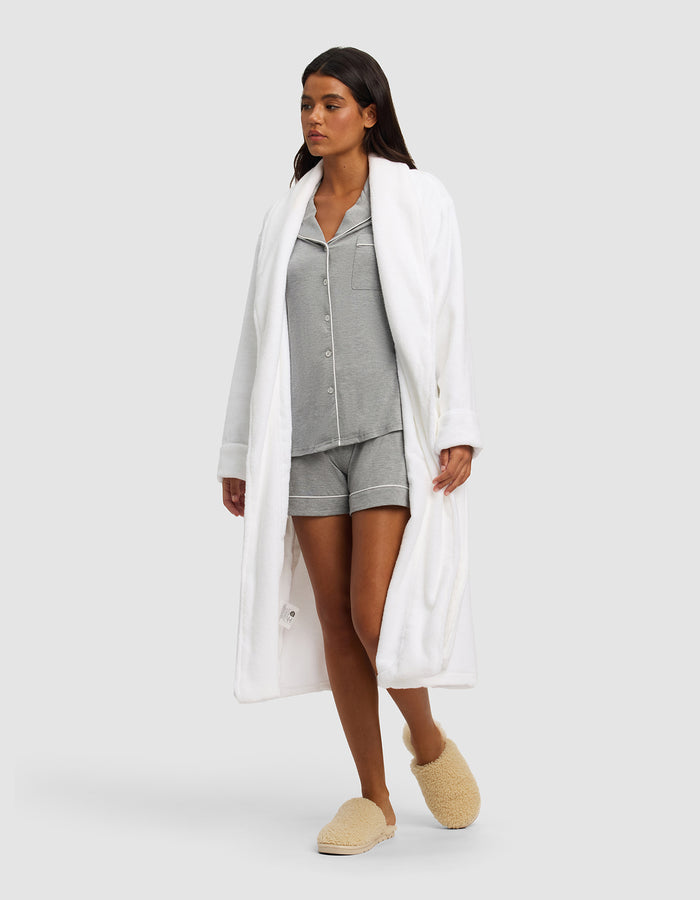 A woman models the Cozy Earth Luxe Bath Robe over gray pajama shorts and matching top, paired with beige fuzzy slippers, against a plain light gray background.|Color:White