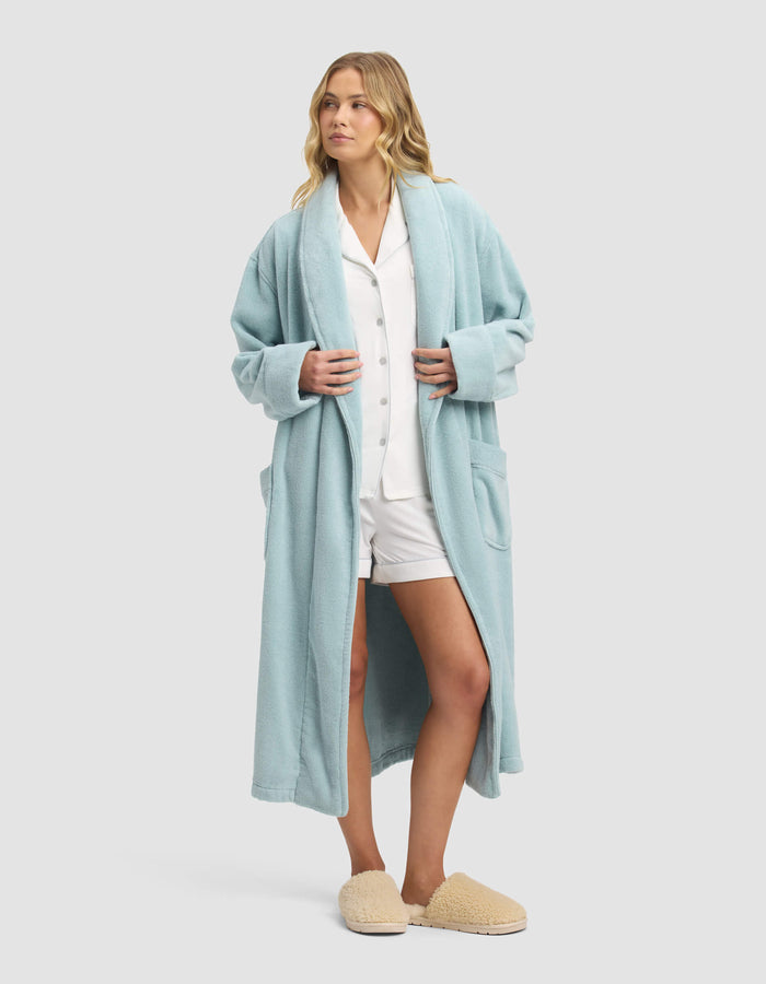 A woman with wavy blonde hair wears the Cozy Earth Luxe Bath Robe over white pajamas and beige slippers, looking relaxed against a plain light gray background.|Color:Tide