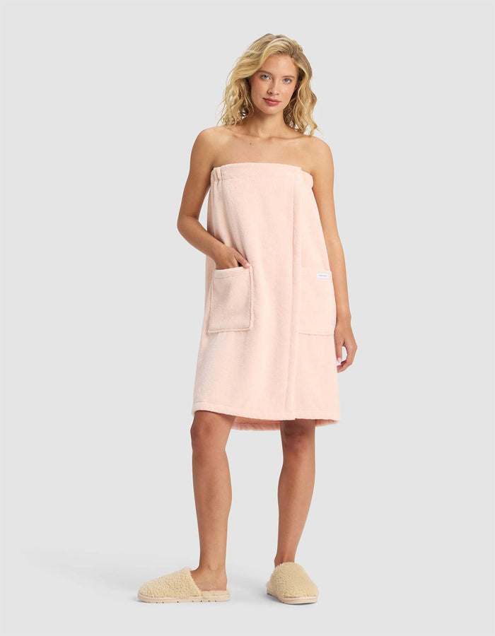 A woman with blonde hair wears the Cozy Earth Luxe Bath Wrap in light pink, featuring two front pockets, along with beige slippers, standing against a plain light gray background.|Color:Blossom