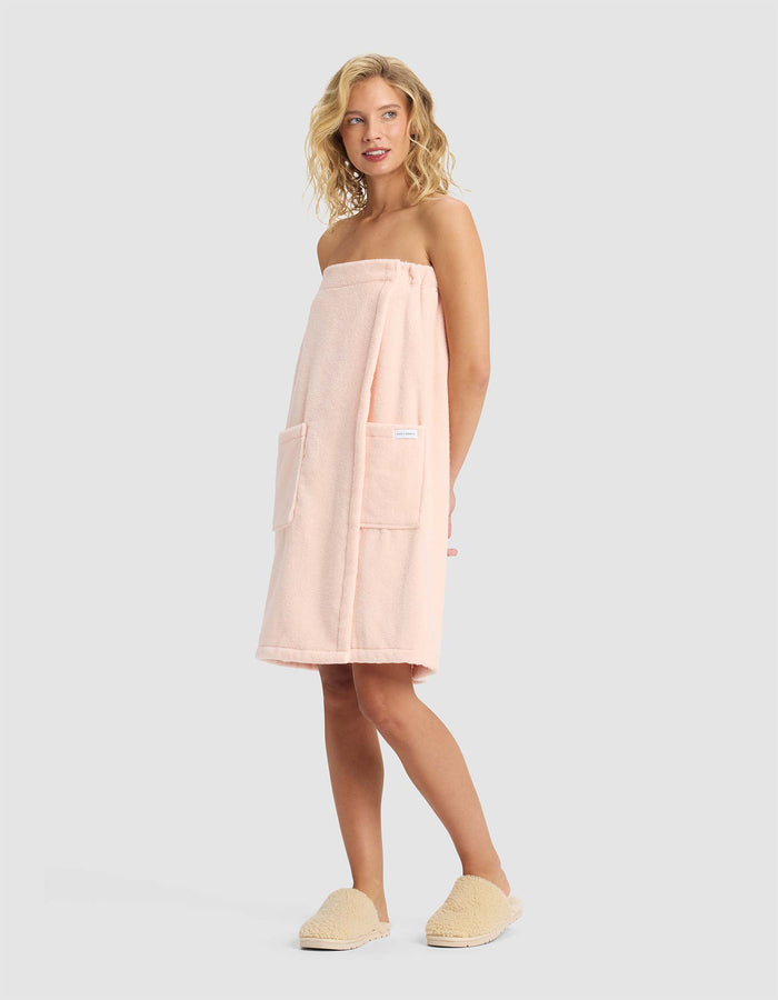 A woman with wavy blonde hair wears the Luxe Bath Wrap by Cozy Earth in pale pink, featuring two large front pockets, along with beige slippers, standing against a plain light gray background.|Color:Blossom