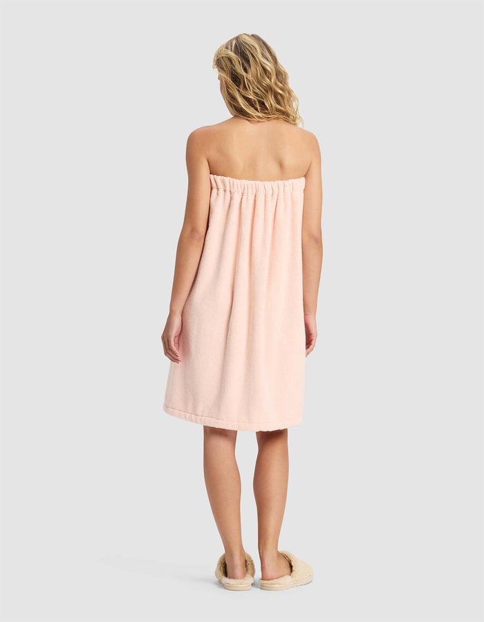 A woman with wavy blonde hair stands facing away, wearing the Cozy Earth Luxe Bath Wrap in soft light pink and beige slippers, set against a plain light gray background.|Color:Blossom