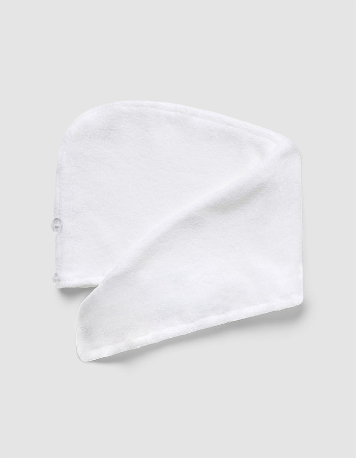 The Luxe Hair Towel by Cozy Earth, made of white microfiber with a button, is folded in a spiral and laid flat on a light gray background.|Color:White