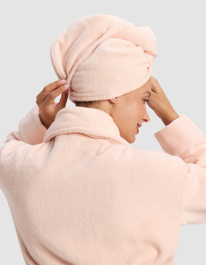 A person adjusts a Luxe Hair Towel by Cozy Earth, wrapped around their head, while wearing a light pink bathrobe, viewed from the back and side.|Color:Blossom