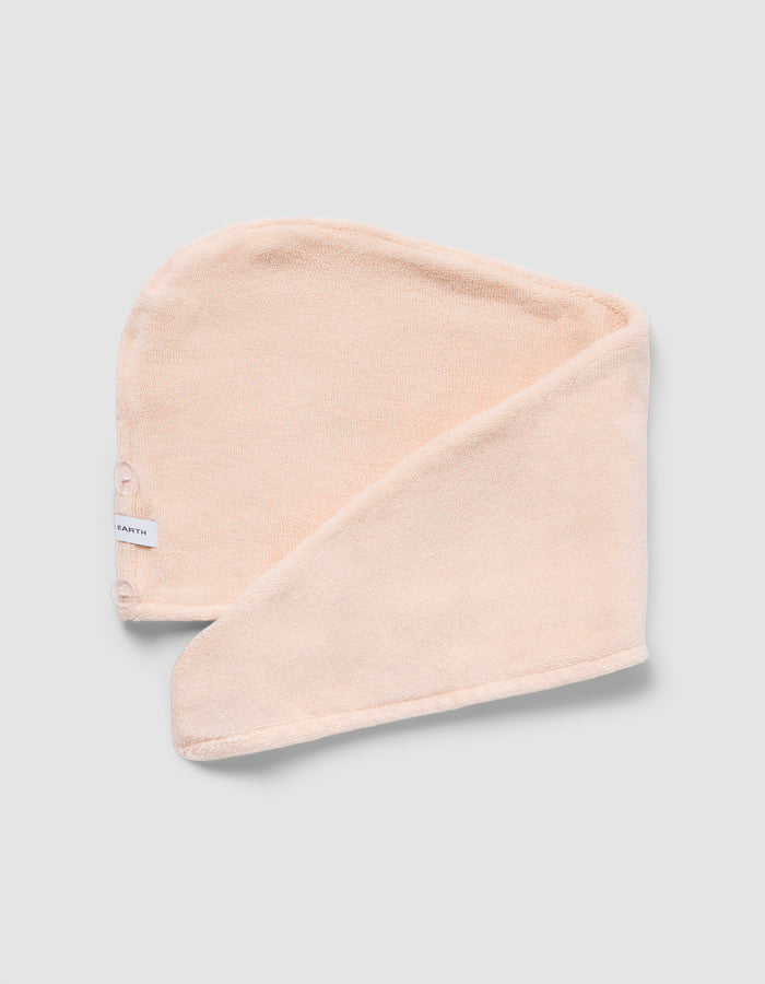 The Luxe Hair Towel by Cozy Earth is a soft, light beige microfiber towel with a white label and two snap buttons. Displayed partially folded on a light gray background, it highlights its rectangular shape and smooth texture.|Color:Blossom