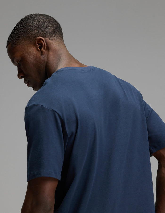 A man with short hair is shown from the back and side, looking downward against a plain gray background while wearing the Cozy Earth Men’s All Day Tee in navy blue.|Color:Eclipse