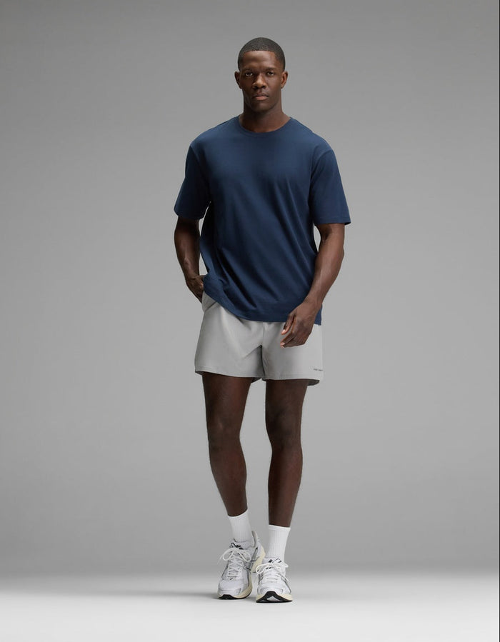 A man stands against a plain gray background wearing the Cozy Earth Men’s All Day Tee in navy blue, paired with light gray shorts, white socks, and white sneakers, with his hands in his pockets.|Color:Eclipse