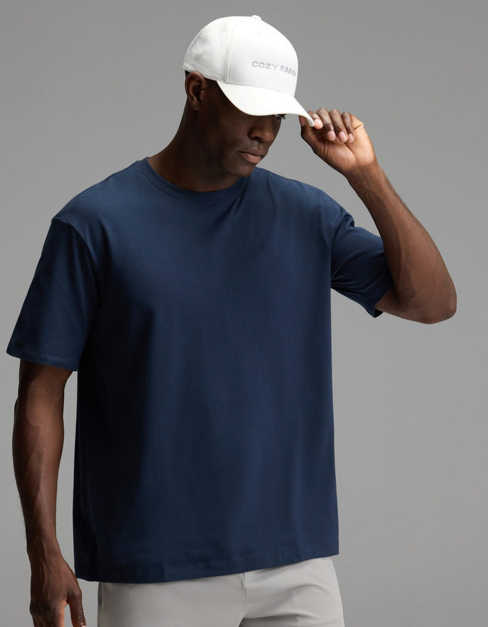 A man wearing the Cozy Earth Men’s All Day Tee in navy blue, paired with light gray pants and a white “COZY FARM” baseball cap, looks down and adjusts his cap against a plain gray background.|Color:Eclipse