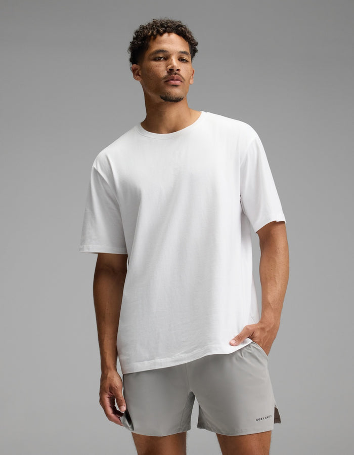 A man stands against a gray background wearing the Cozy Earth Men’s All Day Tee in white and light gray shorts, with one hand in his pocket and a neutral expression.|Color:White