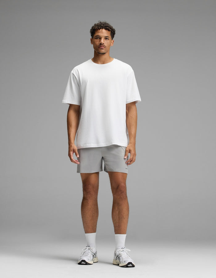 A man stands facing forward against a plain gray background, wearing the Men’s All Day Tee by Cozy Earth, paired with light gray shorts, white socks, and white sneakers.|Color:White