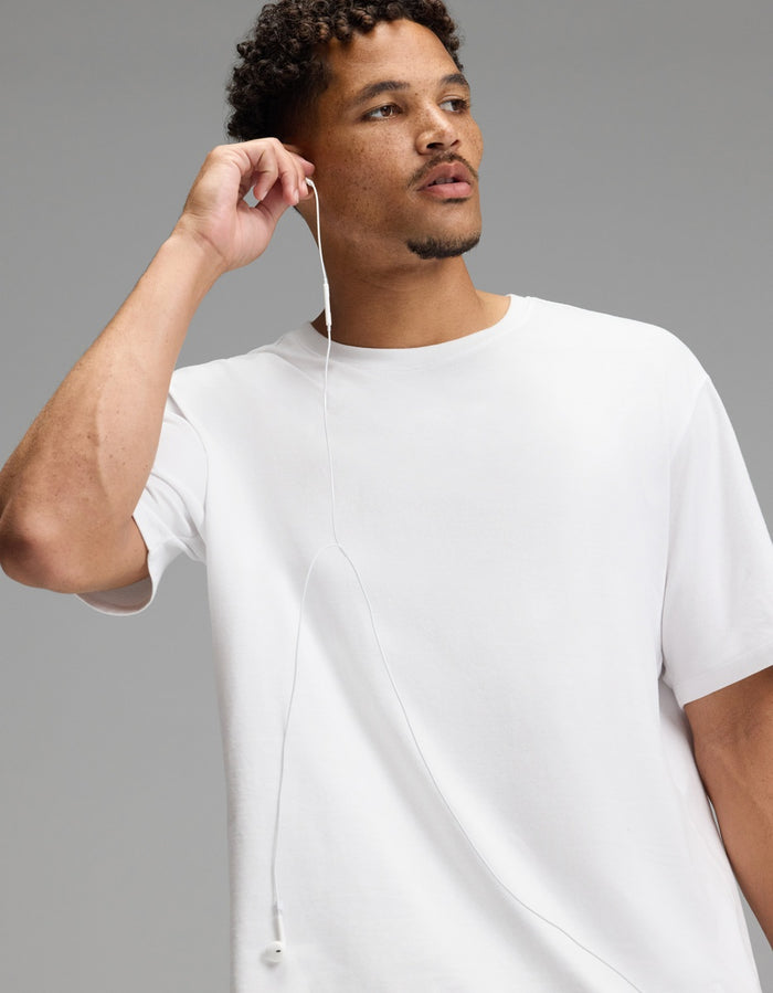 A man wearing the Cozy Earth Men’s All Day Tee in plain white holds an earbud to his ear, gazing off to the side with a neutral expression against a simple gray background.|Color:White