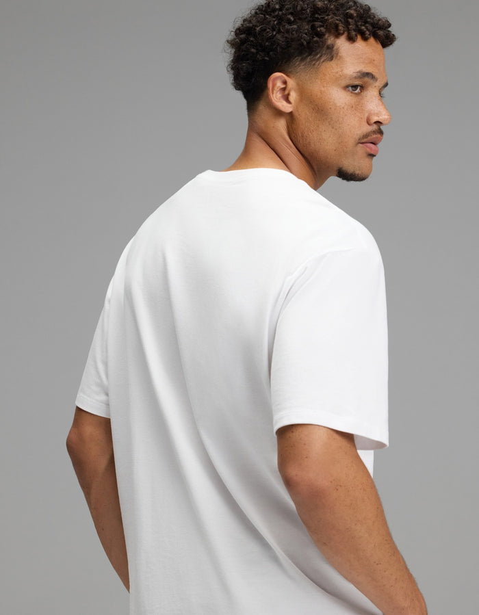 A man with short curly hair and a trimmed beard, wearing the Cozy Earth Men’s All Day Tee in plain white, turns to look over his right shoulder against a neutral gray background.|Color:White
