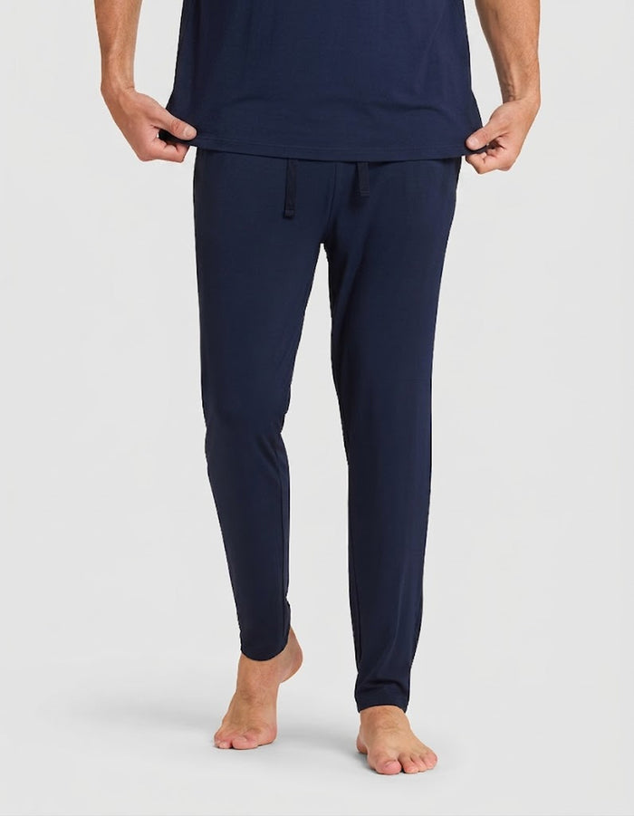 A person stands barefoot in a Cozy Earth Men's Bamboo Stretch-Knit Lounge Tee & Pant Pajama Set in navy blue, holding the shirt slightly lifted against a plain white background.|Color:Navy