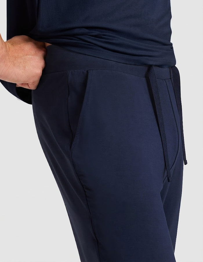 A person wearing Cozy Earth Men's Bamboo Stretch-Knit Lounge Tee & Pant Pajama Set, featuring dark blue pants with a drawstring waistband and their right hand in the pocket, shown from the waist down against a plain background.|Color:Navy