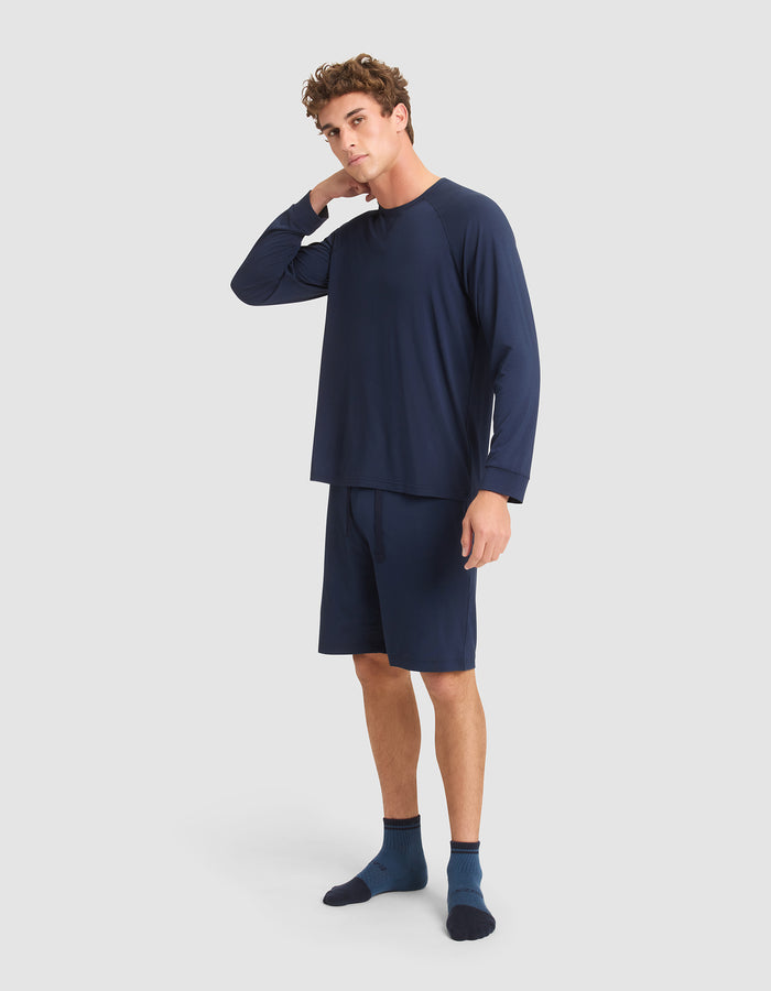 A young man stands against a plain light background, wearing the Cozy Earth Men's Bamboo Stretch-Knit Long Sleeve in navy blue. He touches the back of his neck with one hand and gazes slightly to the side. |Color:Navy