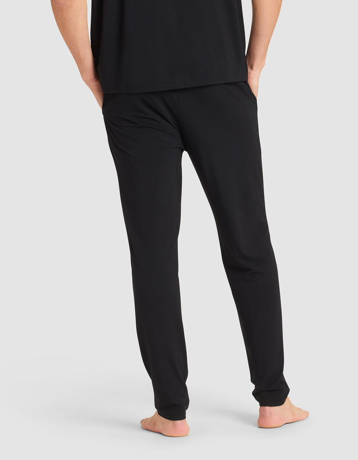 A person shown from the back, barefoot with hands in pockets, wearing Cozy Earth's Men's Bamboo Stretch-Knit Pajama Pant in black and a matching black shirt against a plain light background. |Color:Black
