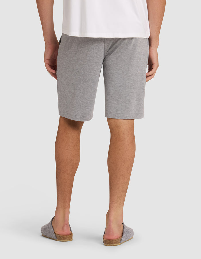 A person wearing Cozy Earth's Men's Bamboo Stretch-Knit Pajama Short in gray, paired with a white shirt and gray slip-on slippers, stands with their back to the camera against a plain background. |Color:Heather Grey