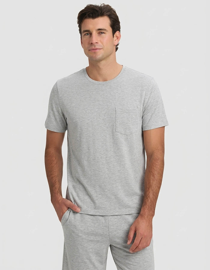 A man models the Cozy Earth Men's Bamboo Stretch-Knit Short Sleeve Pajama Set—light gray with a chest pocket and matching pants—standing with one hand in his pocket against a plain light background.|Color:Heather Grey
