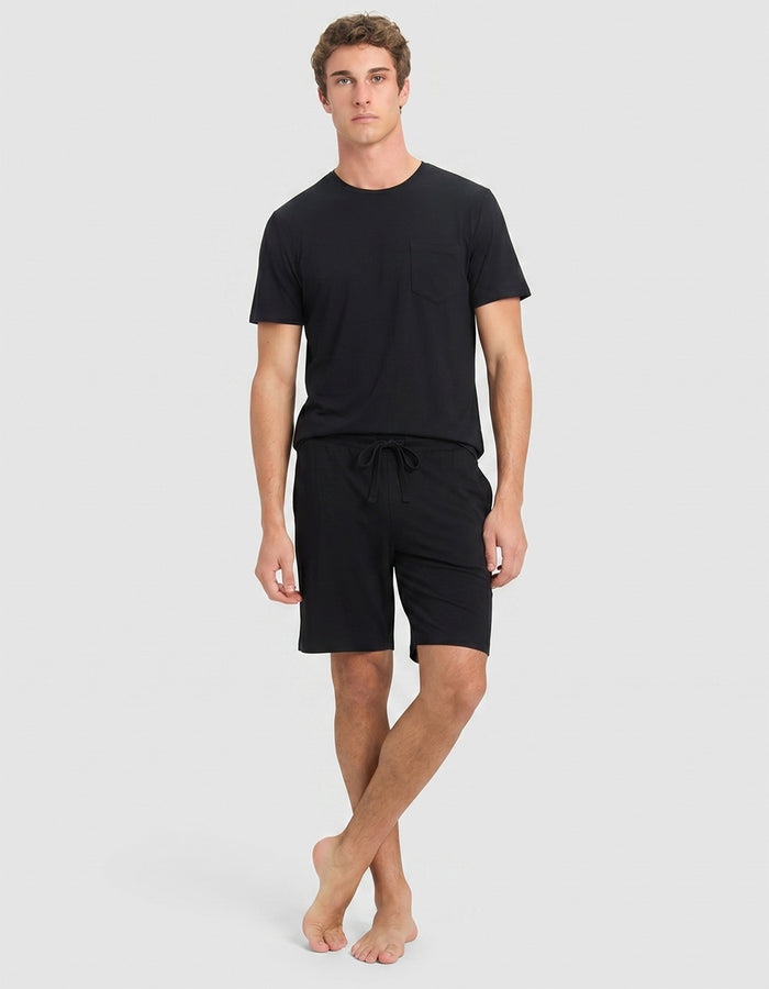 A young man with curly brown hair wears the Cozy Earth Men's Bamboo Stretch-Knit Lounge Tee & Shorts in black. He stands against a light gray background, looking to his left with a neutral expression.|Color:Black