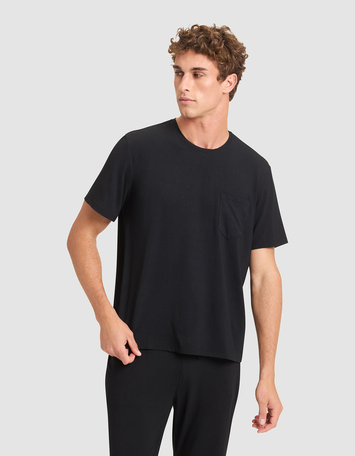 A young man with curly brown hair wears the Cozy Earth Men's Bamboo Stretch-Knit Lounge Tee in black, paired with matching pants. He stands against a light gray background, looking to his left with a neutral expression.|Color:Black