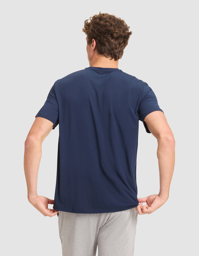 A man with curly hair faces away from the camera, wearing Cozy Earth's Men's Bamboo Stretch-Knit Lounge Tee in navy blue and light gray shorts, holding the hem of his shirt with both hands against a plain background.  |Color:Navy