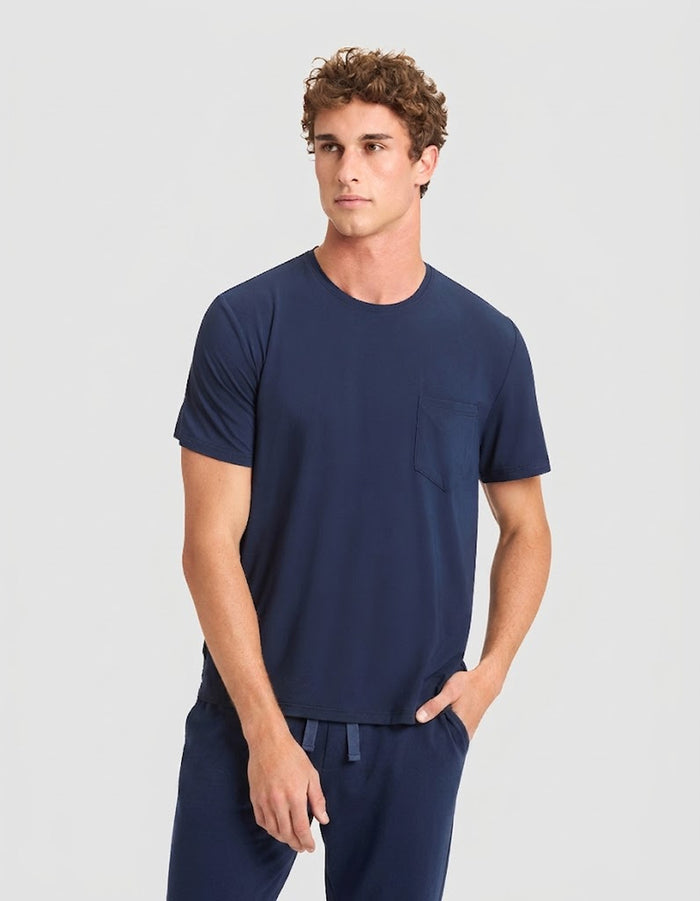 A young man with short curly hair models the Cozy Earth Men's Bamboo Stretch-Knit Lounge Tee & Pant Pajama Set in navy, featuring a short-sleeve chest-pocket tee and matching drawstring pants, against a plain light gray background.|Color:Navy