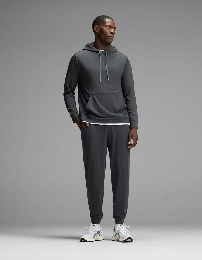 A man stands against a grey background, wearing the Cozy Earth Men’s Brushed Bamboo Hoodie & Jogger Set in Charcoal, paired with white athletic shoes. He looks to the side with his hands by his sides.|Color:Charcoal