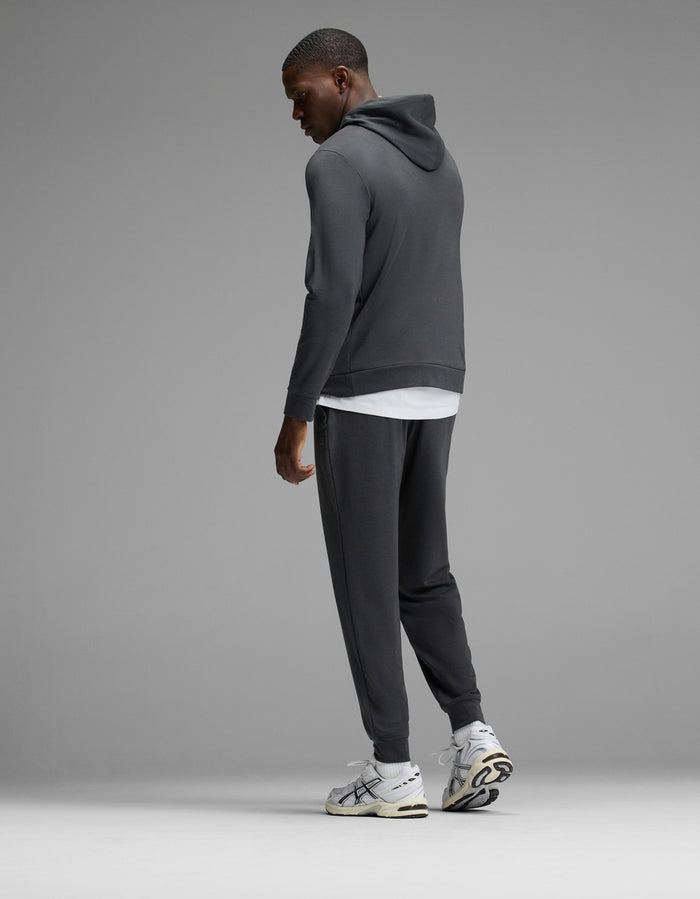 A man wearing the Cozy Earth Men’s Brushed Bamboo Hoodie & Jogger Set in dark gray, paired with a white t-shirt and white sneakers, stands facing away and looks over his shoulder against a plain gray background.|Color:Charcoal
