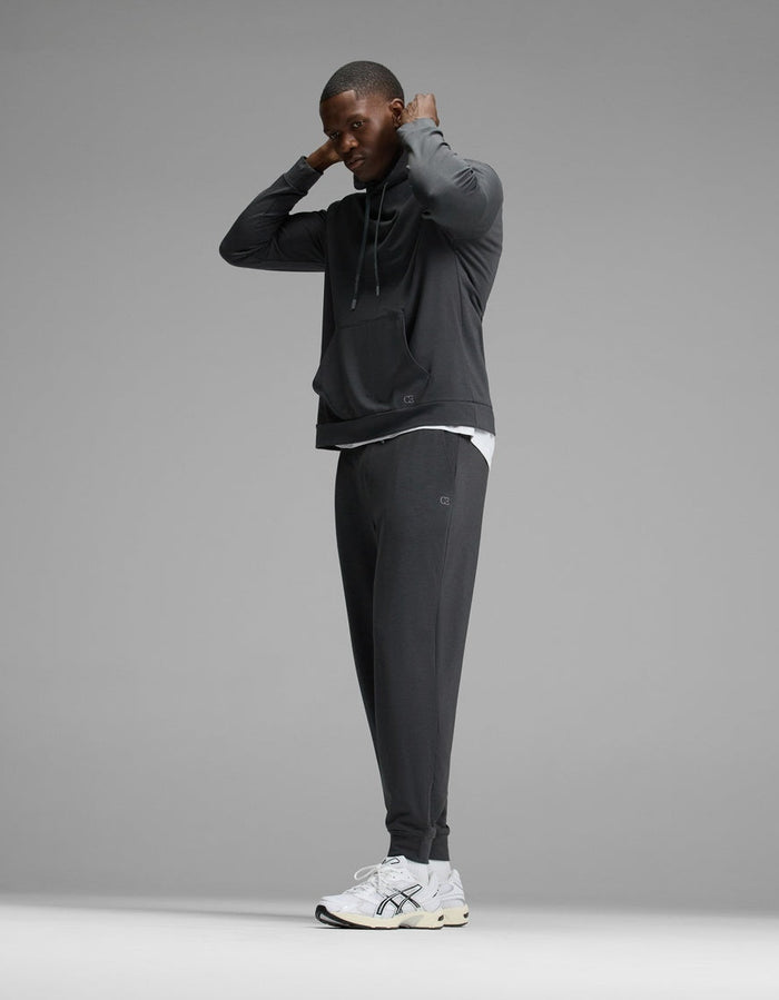 A man wearing the Cozy Earth Men’s Brushed Bamboo Hoodie & Jogger Set stands adjusting his hood, paired with white sneakers. The background is plain gray.|Color:Charcoal