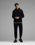 A man stands against a gray background, wearing the Cozy Earth Men’s Brushed Bamboo Hoodie & Jogger Set in black, with a white T-shirt visible beneath the hoodie and black sneakers. He looks slightly to the side, hands together.|Color:Black