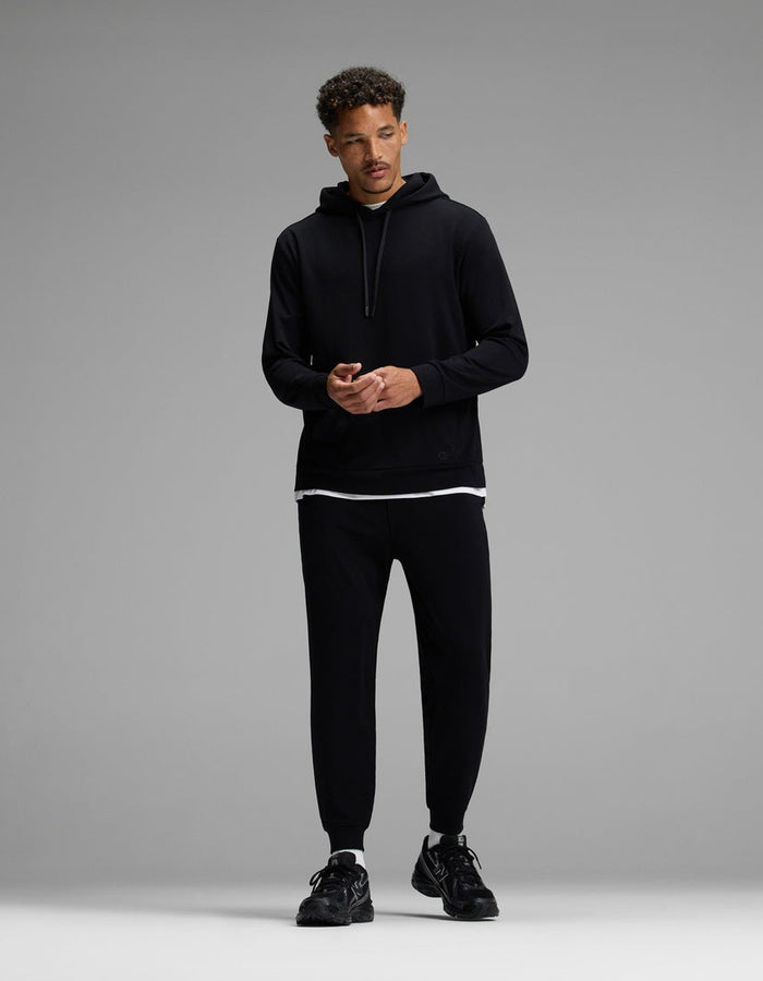 A man stands against a gray background, wearing the Cozy Earth Men’s Brushed Bamboo Hoodie & Jogger Set in black, with a white T-shirt visible beneath the hoodie and black sneakers. He looks slightly to the side, hands together.|Color:Black
