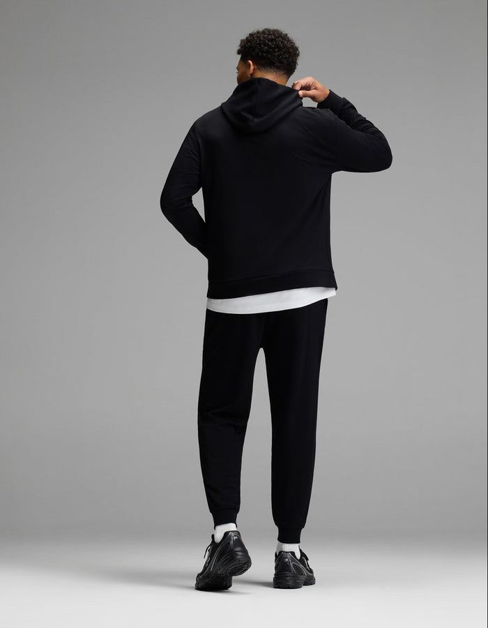 A person stands facing away, dressed in the Cozy Earth Men’s Brushed Bamboo Hoodie & Jogger Set in black, paired with black sneakers and a white shirt peeking out underneath. The background is plain gray.|Color:Black