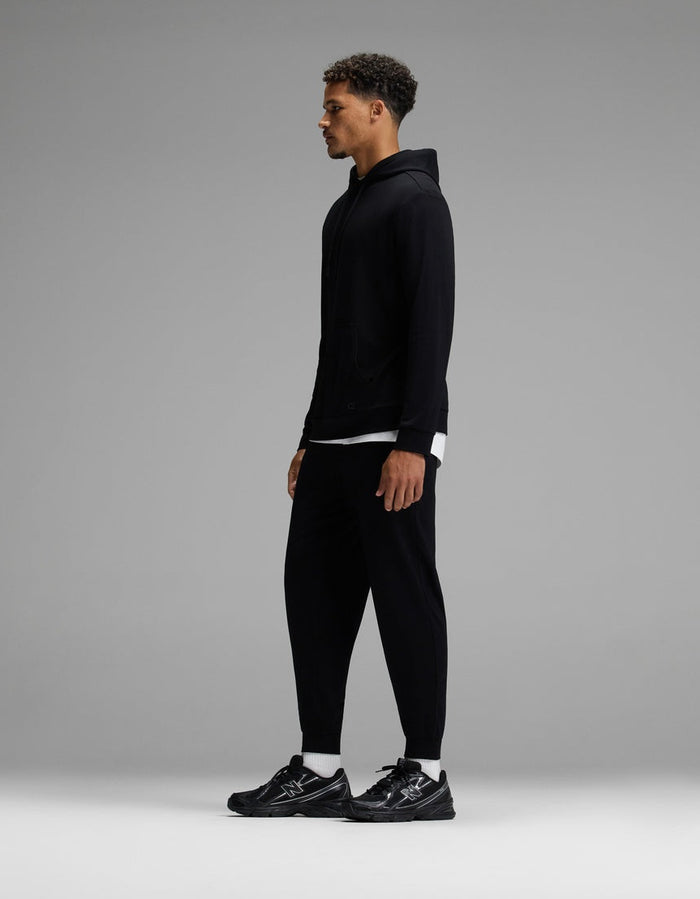 A man stands in profile against a plain gray background, wearing the Cozy Earth Men’s Brushed Bamboo Hoodie & Jogger Set in black, paired with black sneakers and a white shirt peeking out from under the hoodie.|Color:Black