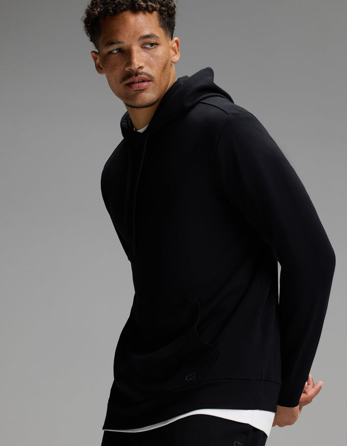 A man with short curly hair models the Cozy Earth Men’s Brushed Bamboo Hoodie & Jogger Set in black over a white shirt, posing against a plain gray background.|Color:Black