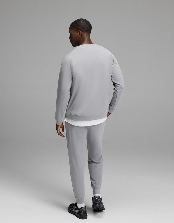 A man stands facing away, wearing the Cozy Earth Men’s Brushed Bamboo Jogger Set in light gray paired with black sneakers, his left hand resting on his hip against a plain, neutral gray background.|Color:Stone