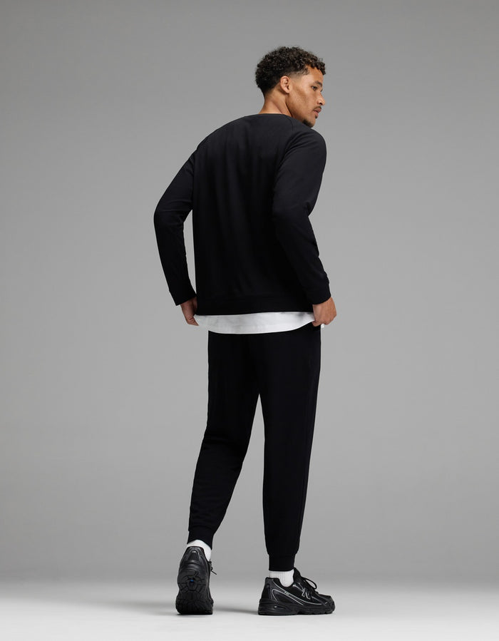A man stands against a gray background, facing away and looking over his shoulder. He wears the Men’s Brushed Bamboo Jogger Set by Cozy Earth, featuring a black long-sleeve top and matching black joggers layered over a white shirt, paired with white socks and black sneakers.|Color:Black