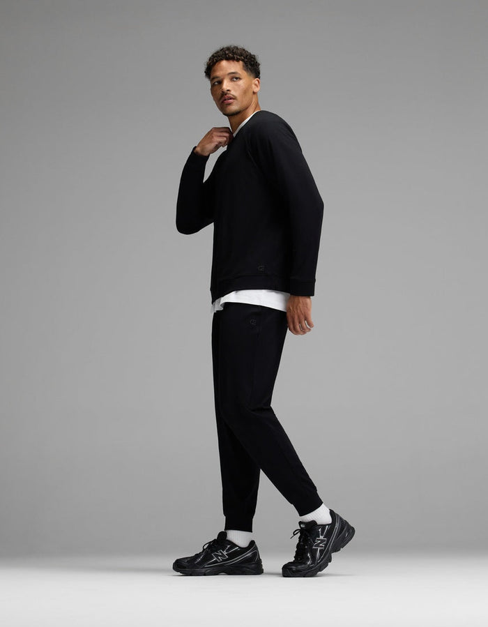 A man stands in a studio wearing the Cozy Earth Men’s Brushed Bamboo Jogger Set in black, paired with black sneakers and a white undershirt. He looks to the side with one hand near his neck, against a plain gray background.|Color:Black