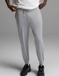 A person is shown from the shoulders down, wearing Cozy Earth’s Men’s Brushed Bamboo Joggers in light gray, paired with a white shirt and black athletic shoes, standing against a plain gray background.|Color:Stone
