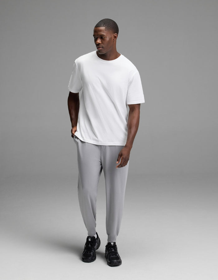 A man wearing Cozy Earth Men’s Brushed Bamboo Joggers in light gray, paired with a plain white T-shirt and black sneakers, stands against a simple gray background, looking slightly down and to the side with one hand in his pocket.|Color:Stone