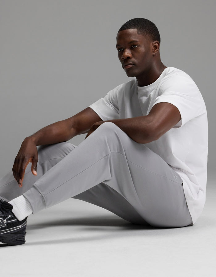A man wearing Cozy Earth’s Men’s Brushed Bamboo Joggers in light gray, paired with a white t-shirt, white socks, and black sneakers, sits on the floor against a plain gray background.|Color:Stone