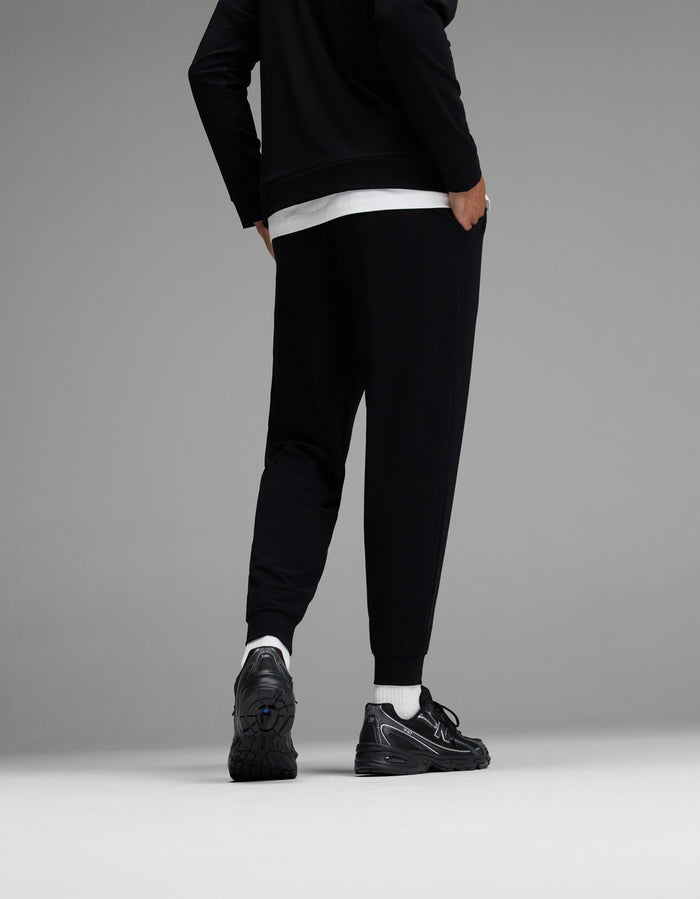 A person stands facing away, wearing black jogger pants, a black jacket, a white shirt, white socks, and black sneakers against a gray background.|Color:Black