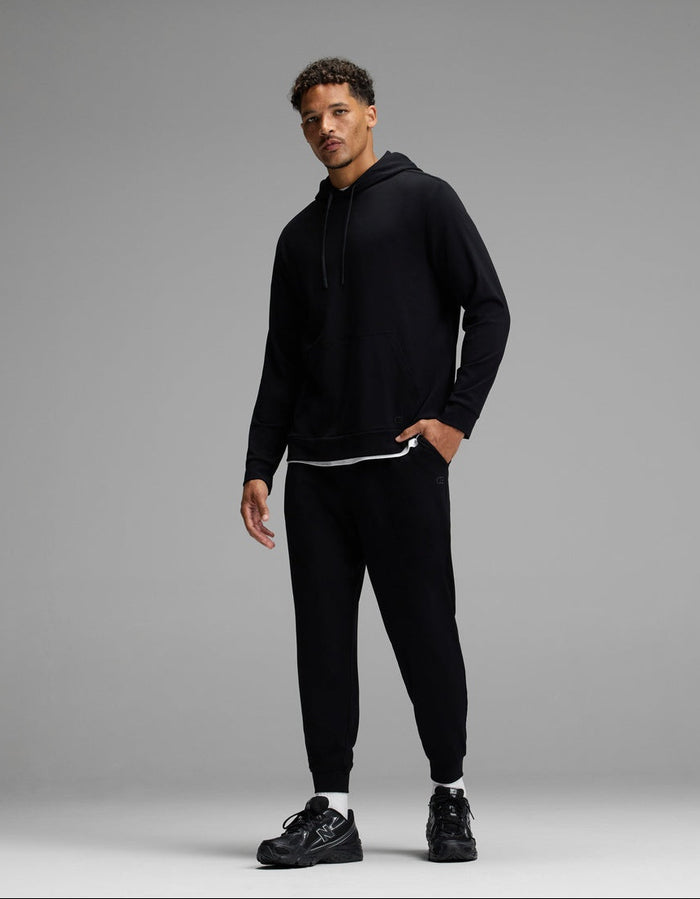 A man stands against a gray background wearing a black hoodie, black jogger pants, and black sneakers. He has short curly hair and is looking at the camera with a relaxed posture.|Color:Black