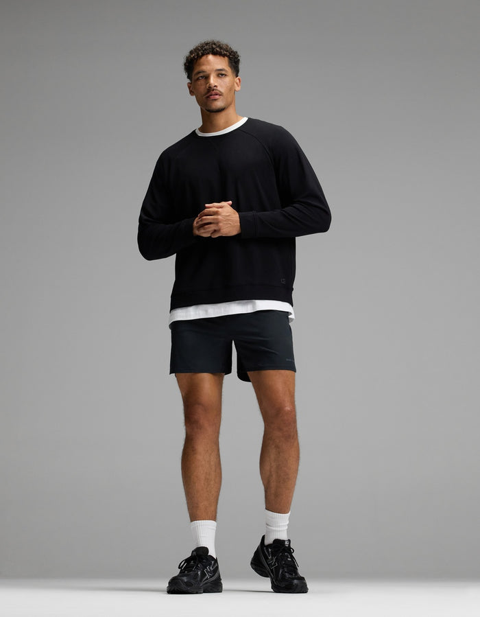 A man stands against a gray background wearing the Cozy Earth Men’s Brushed Bamboo Pullover Crew, paired with black shorts, white crew socks, and black sneakers. He has short curly hair and his hands are clasped in front of him.|Color:Black